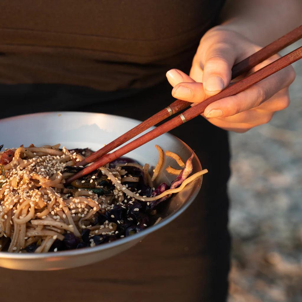 Hand holding wooden chopsticks above a bowl of noodles with sesame seeds outdoors