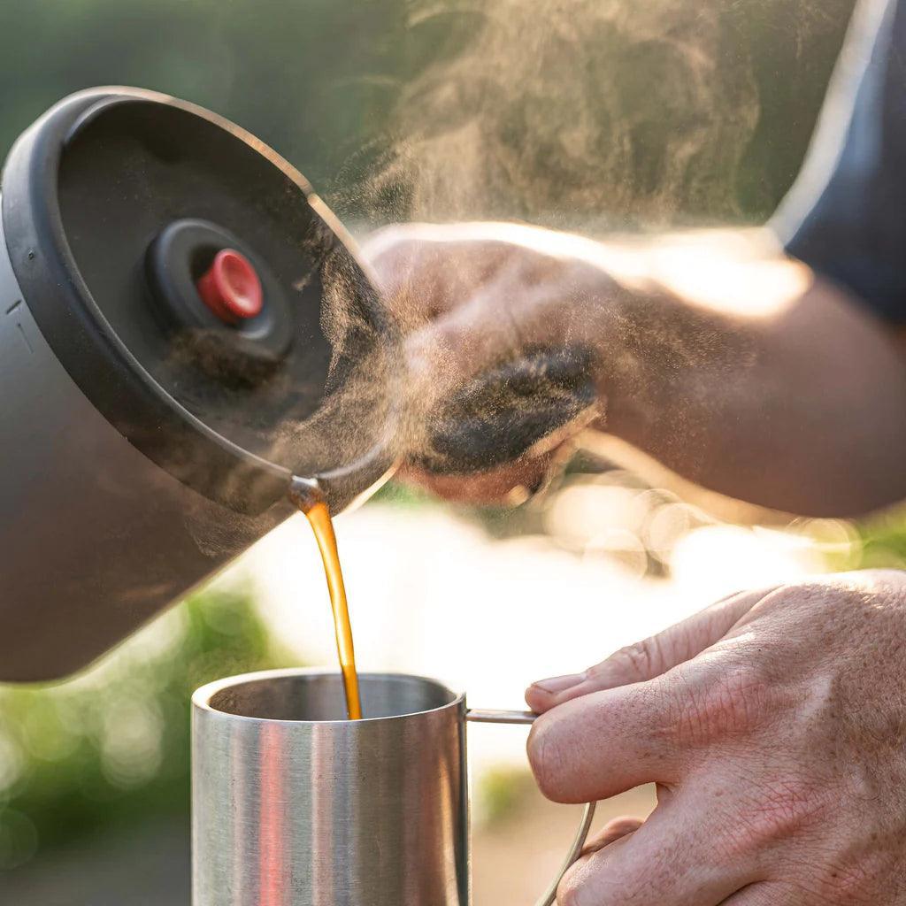 Pouring hot coffee from a Primus campsite press into a metal mug outdoors