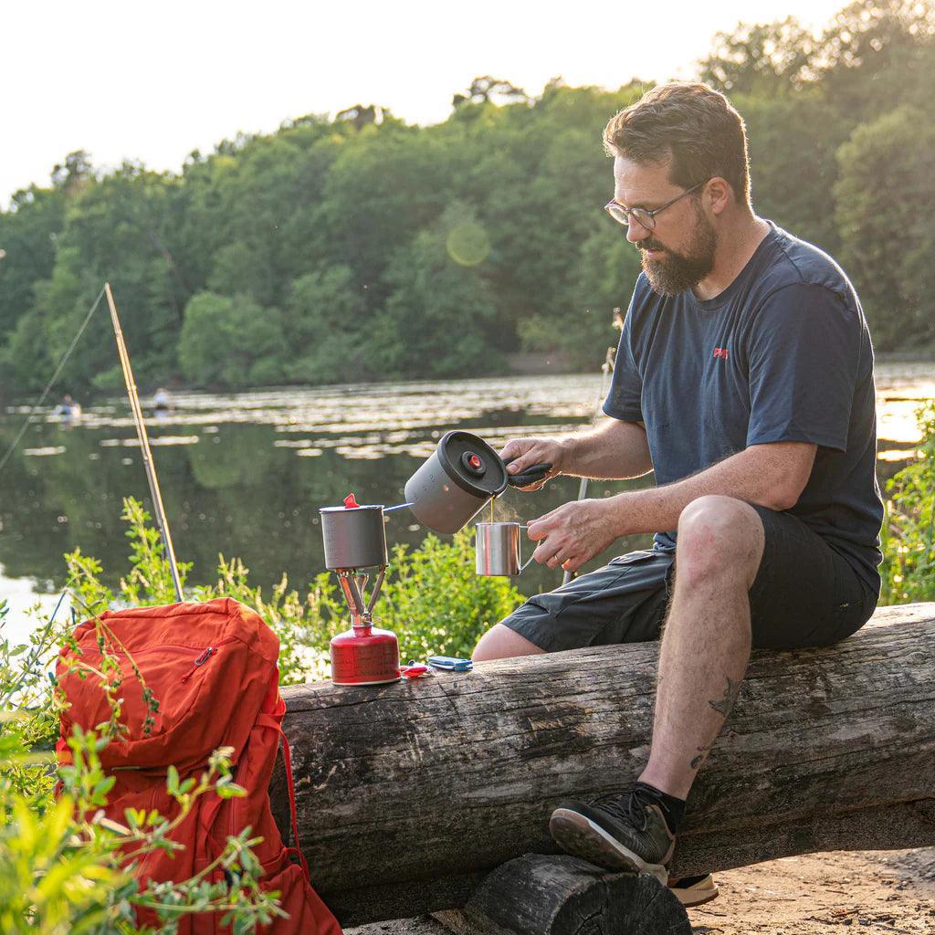 Man making coffee with Primus campsite press on a log by a lake, orange backpack nearby