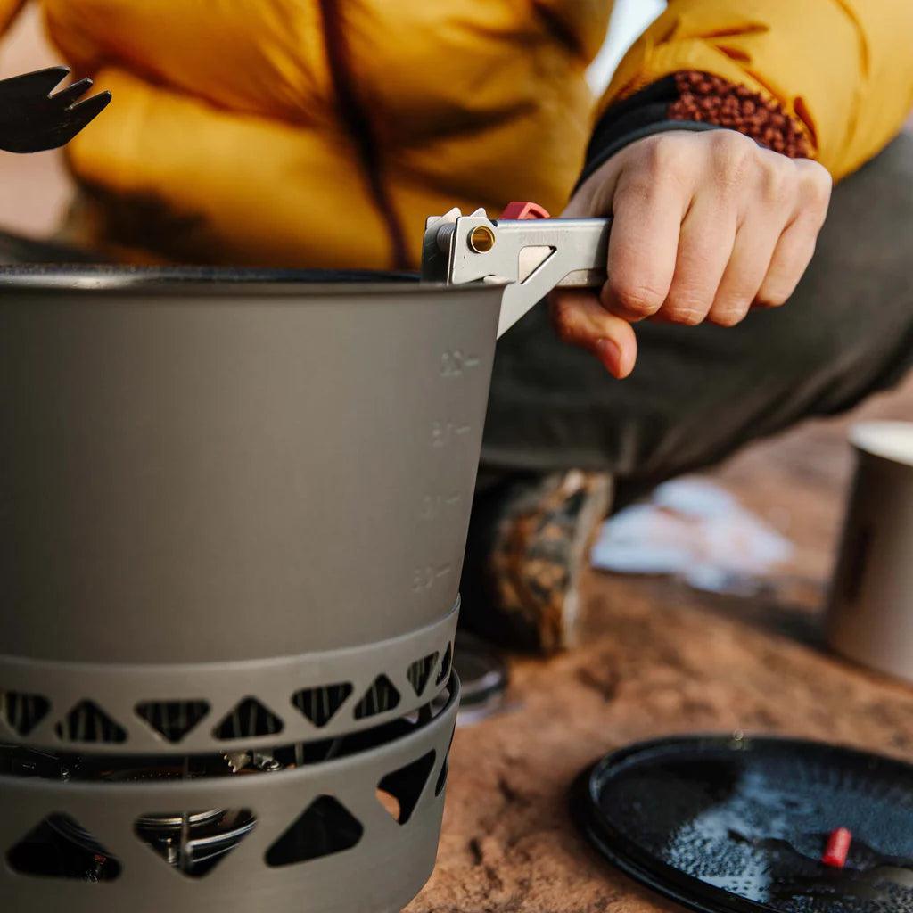 Hand using Primus campsite pot gripper with outdoor camping cookware on a sandy surface