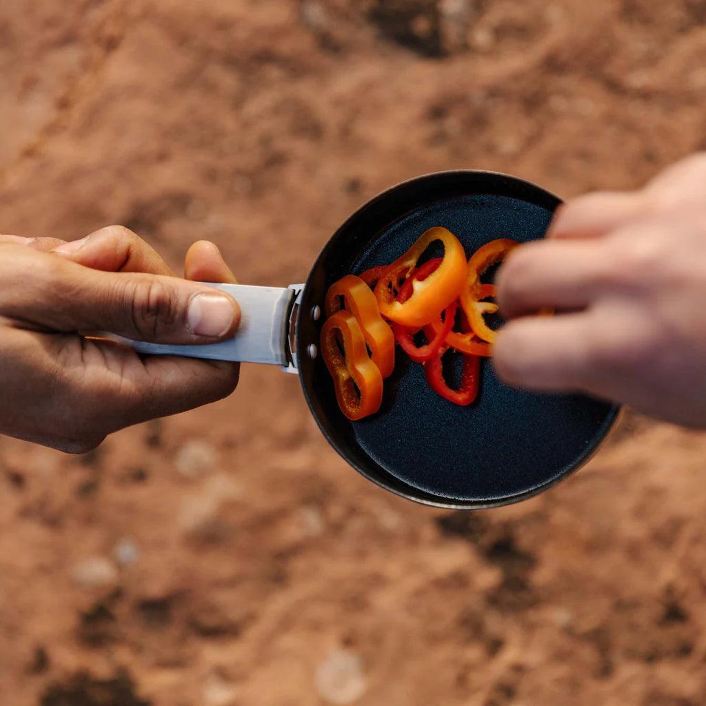 Sliced bell peppers cooking in a small camping pan outdoors on rocky ground
