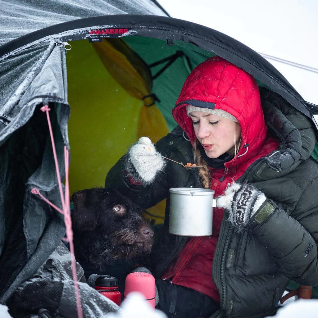 Woman in red hood eating with camping cookware inside a tent in snowy winter with a dog