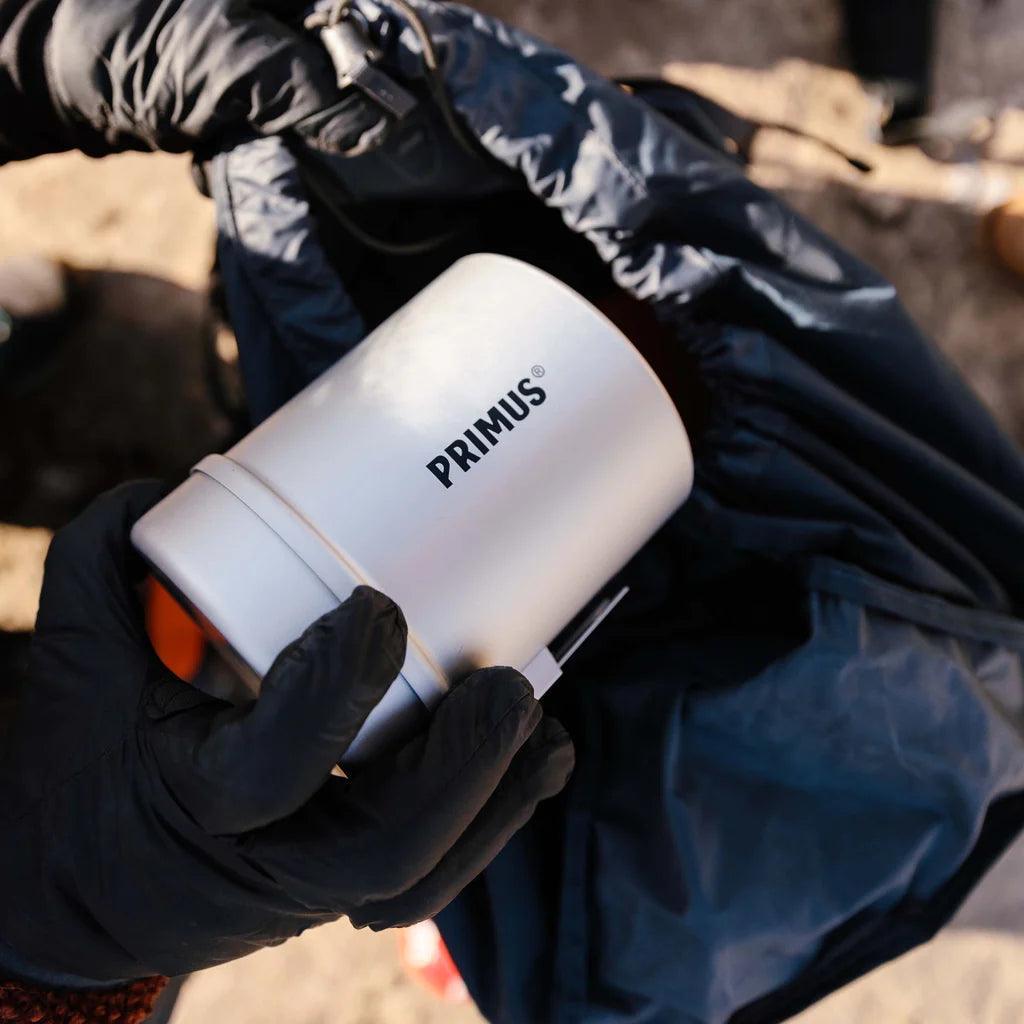 Primus camping cookware kit held by gloved hand over a backpack outdoors