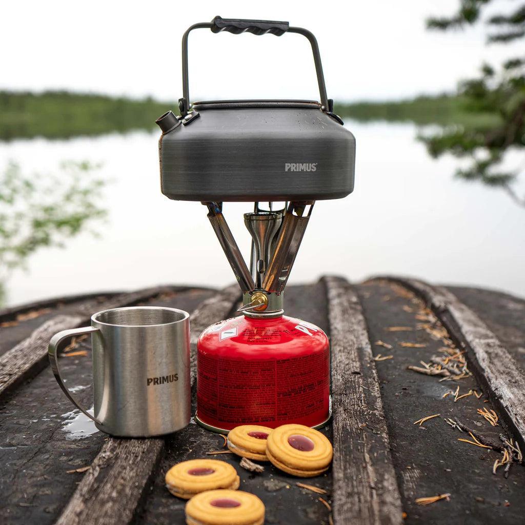 Primus backpacking stove with kettle, metal mug, and cookies on outdoor wooden table by lake
