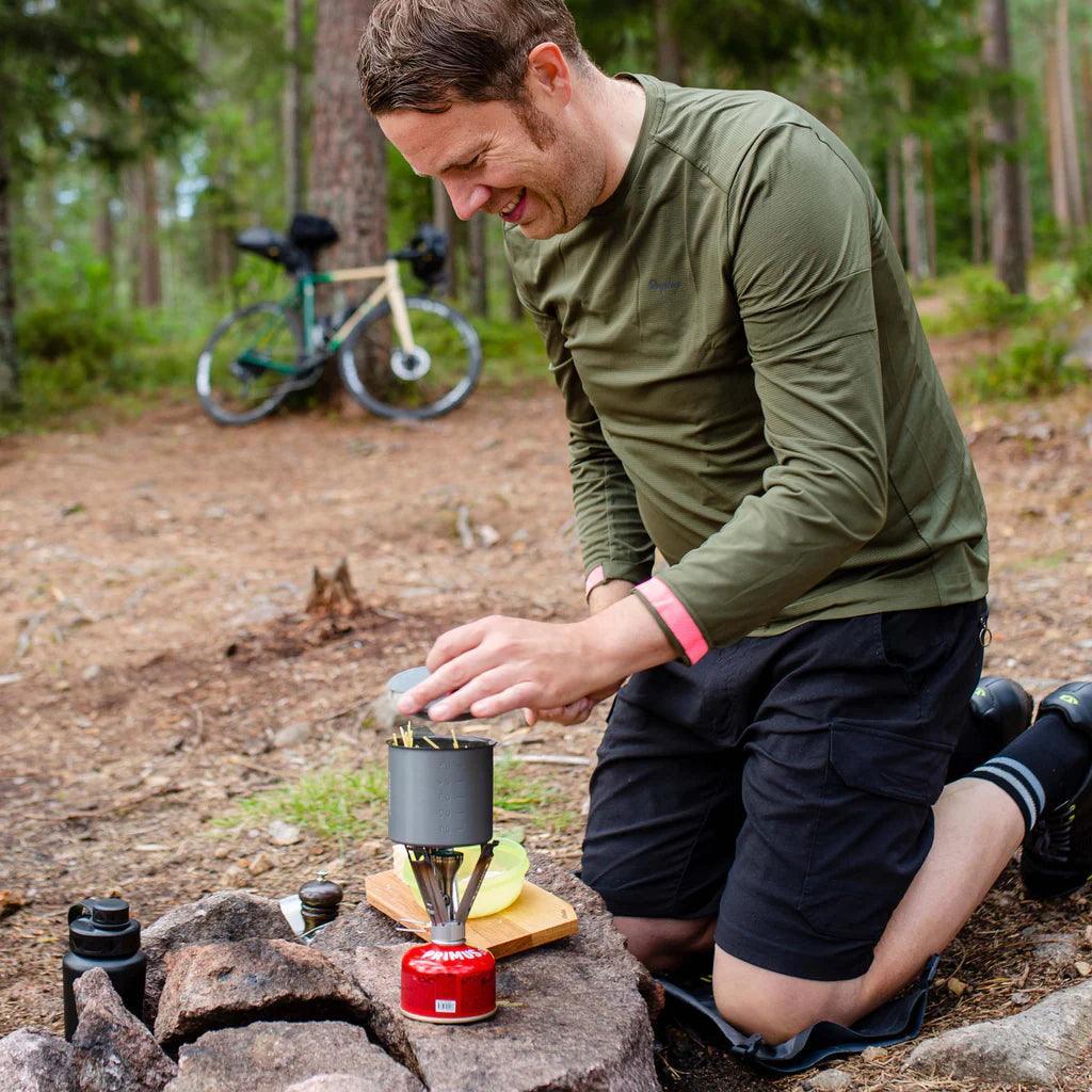 Man cooking with Primus Firestick backpacking stove at forest campsite, bike in background