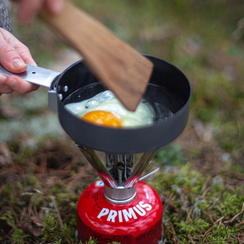 Primus backpacking stove cooking egg outdoors on mossy ground, camping cookware scene