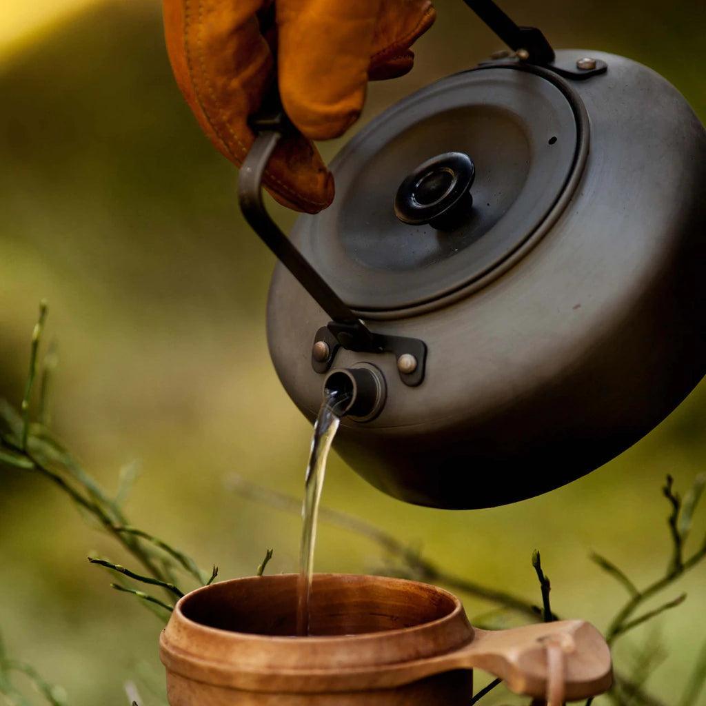 Camp kettle pouring hot water into wooden mug outdoors, camping coffee tea gear
