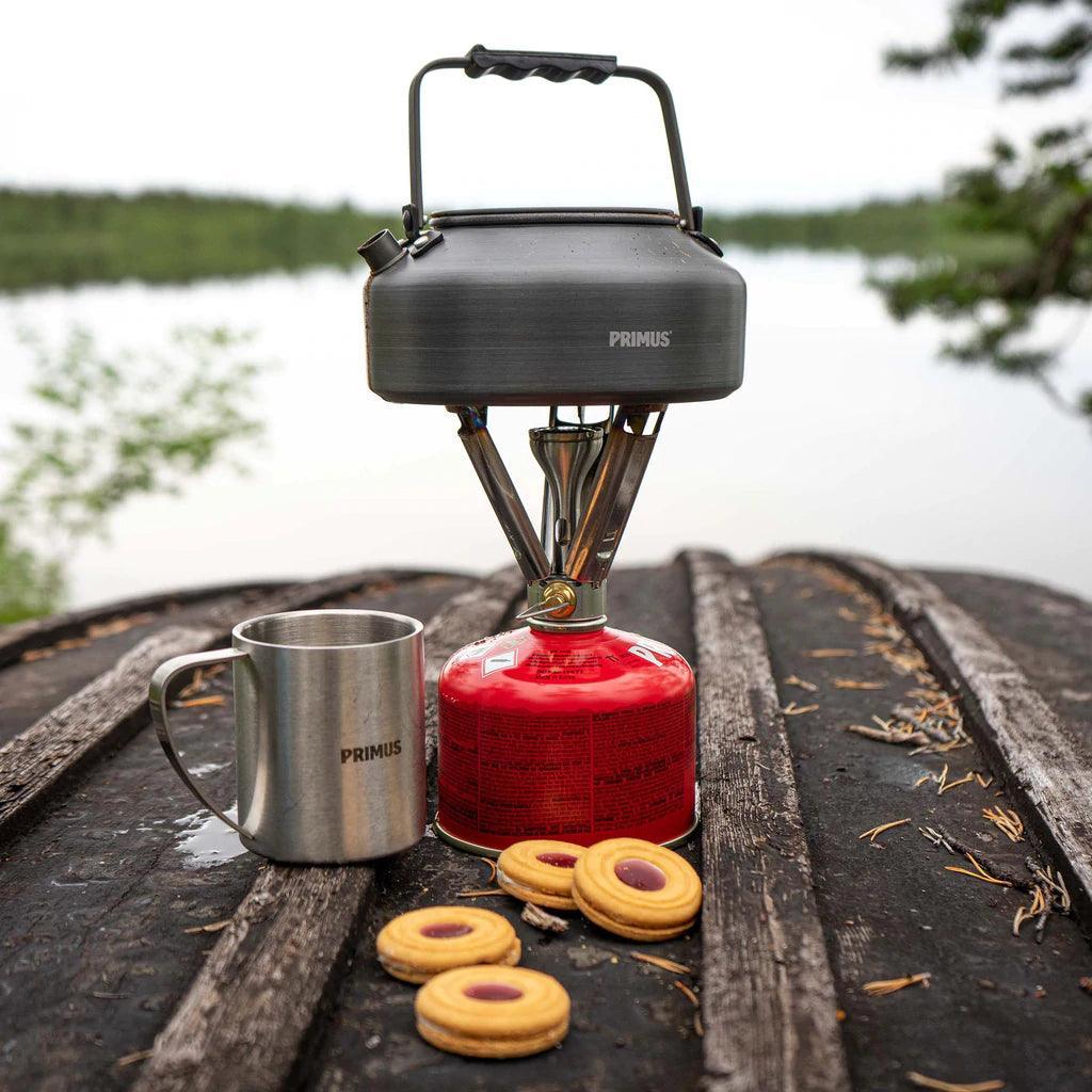 Primus camping kettle on portable gas stove with mug and cookies outdoors by a lake