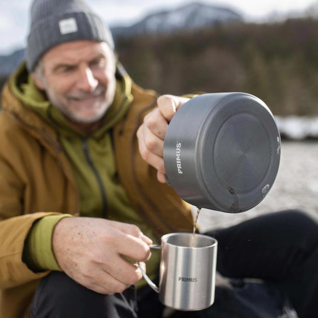 Man pouring coffee from Primus Litech kettle into metal mug outdoors, campsite setting