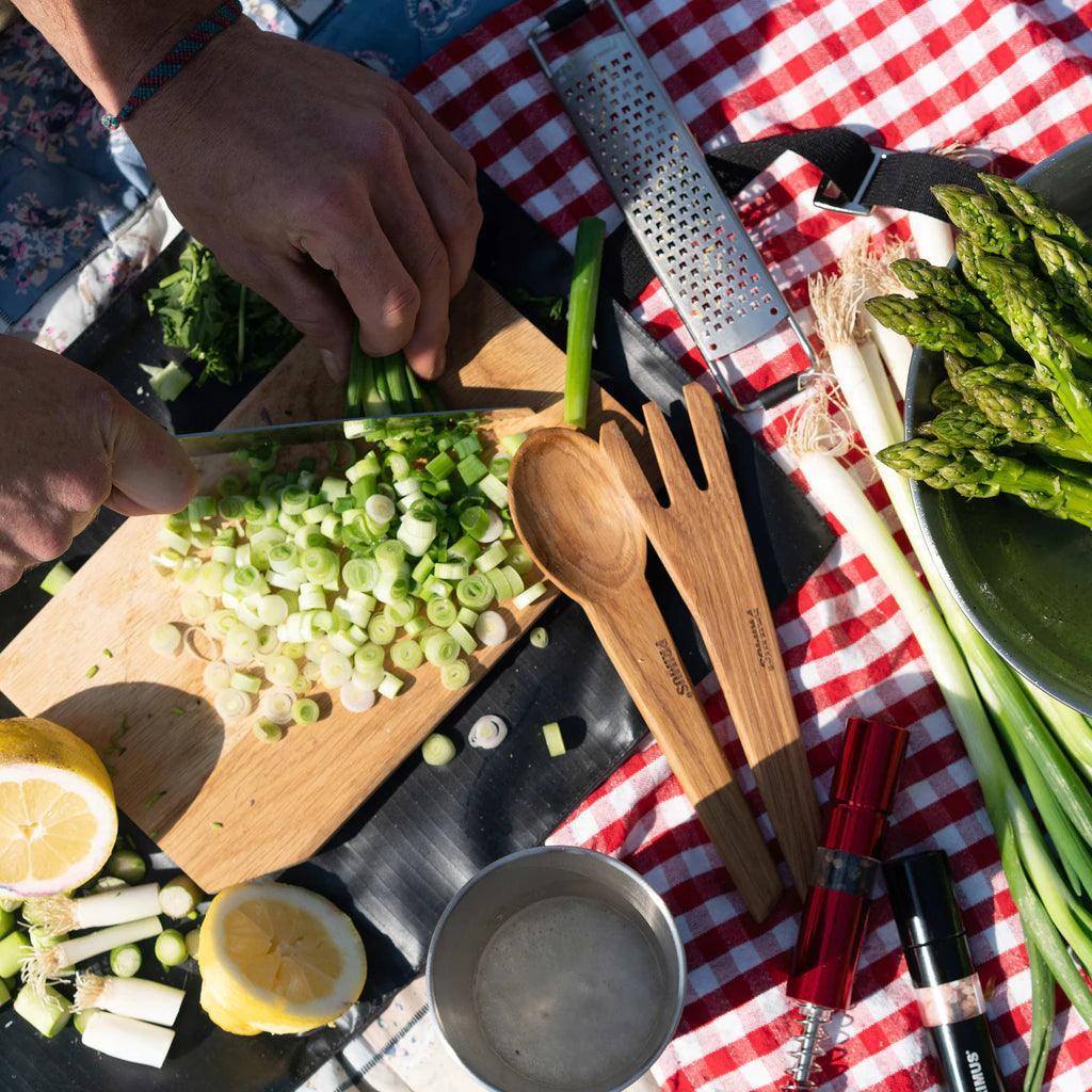 Chopping green onions on a wooden board with Primus campsite salt and pepper mills, asparagus, lemon, and wooden utensils on a picnic blanket.