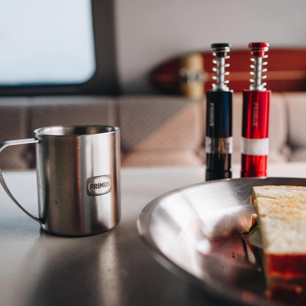 Primus stainless steel camping mug, salt and pepper mills, and sandwich on metal plate indoors