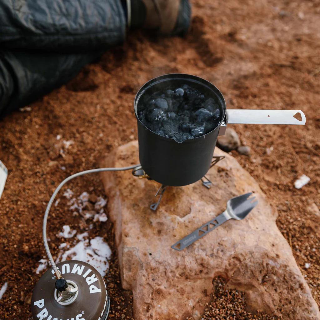 Primus camp stove boiling water in pot with metal spork on rocky ground outdoors
