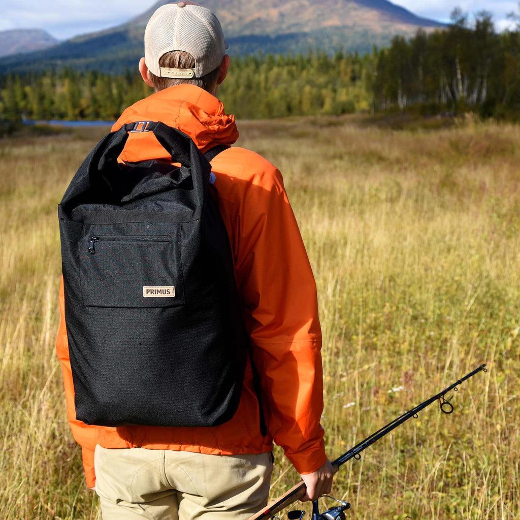 Person in orange jacket with Primus cooler backpack and fishing rod in outdoor field