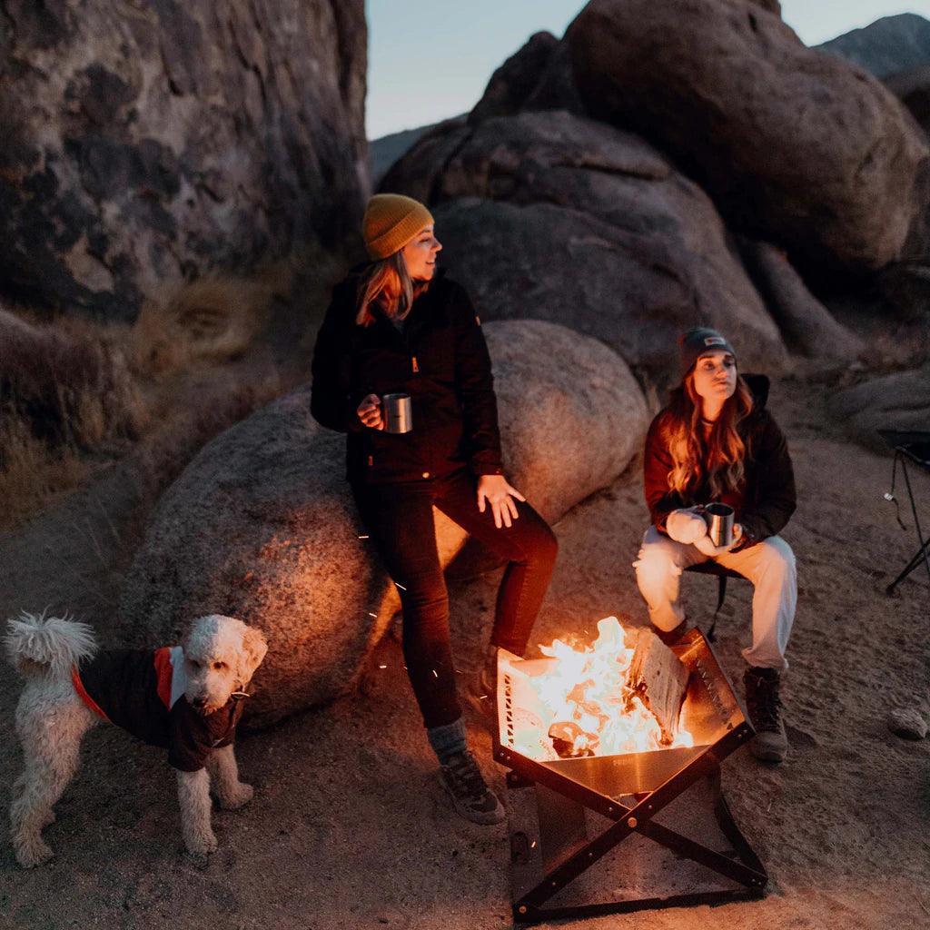 Two women and a dog by Primus Kamoto OpenFire pit outdoors among large rocks at dusk