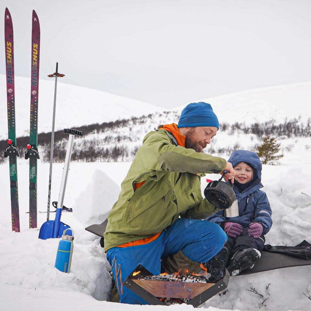 Man and child using Primus Kamoto open fire pit outdoors in snowy winter landscape with skis