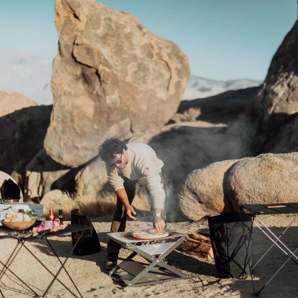 Man grilling on Primus Kamoto open fire pit outdoors with rocks and camping gear visible