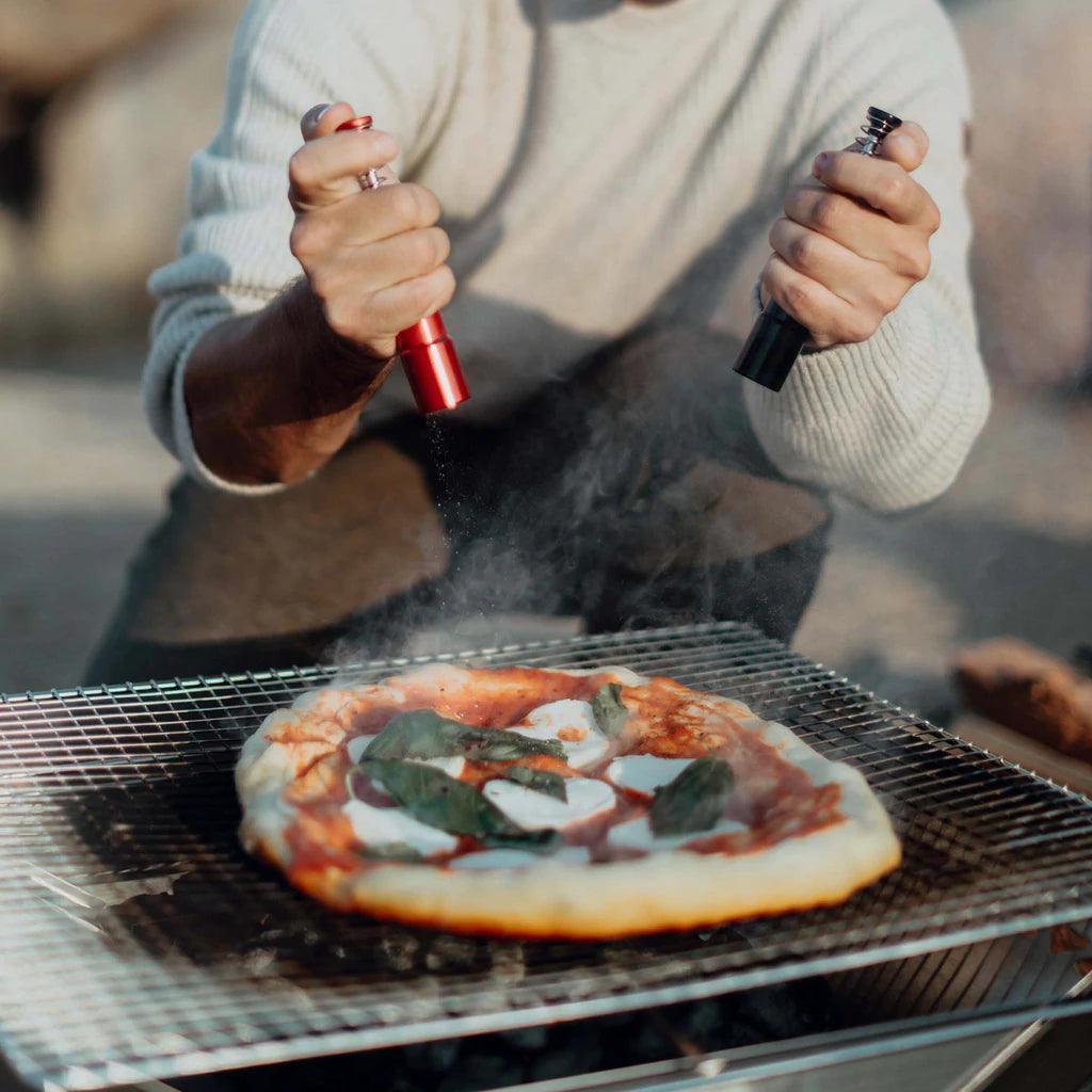Grilling pizza outdoors on a Primus Kamoto open fire pit with person seasoning