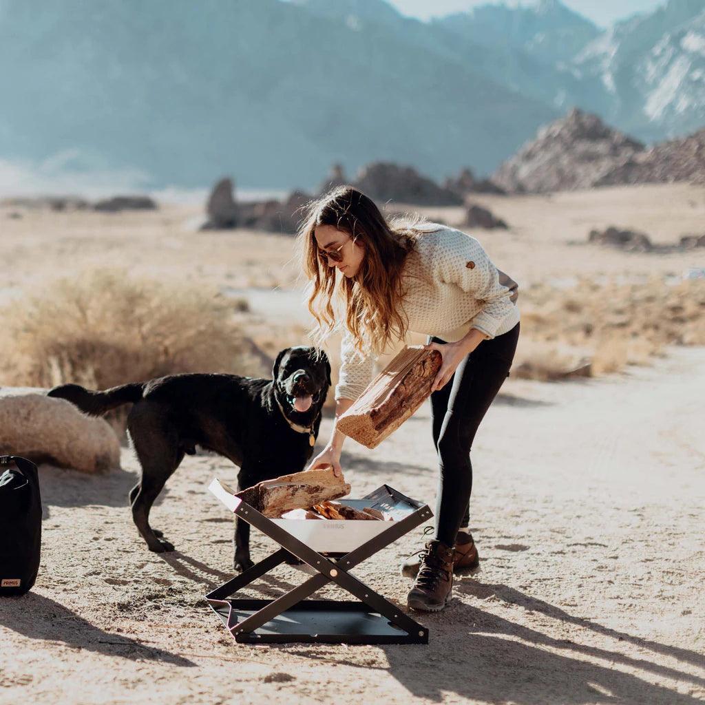 Woman adds firewood to Primus Kamoto open fire pit outdoors with dog, desert mountains background