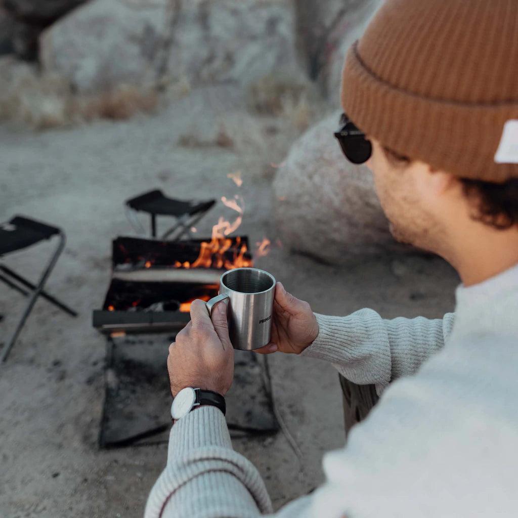 Camper holding metal mug by Primus Kamoto open fire pit outdoors