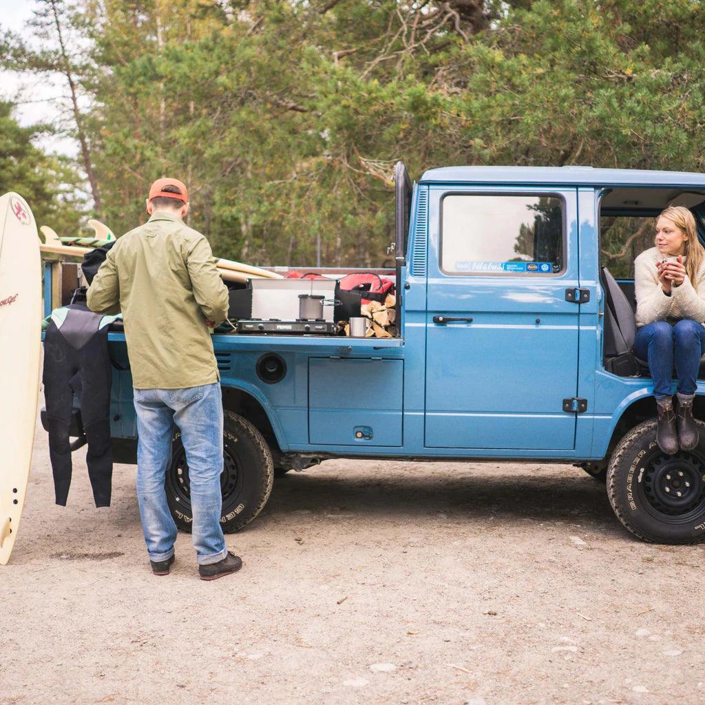 Primus Kinjia camping stove on blue truck tailgate, man prepping gear, woman seated with mug