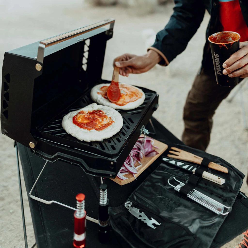 Person spreading sauce on pizza dough using Primus Kuchoma portable gas camp grill outdoors