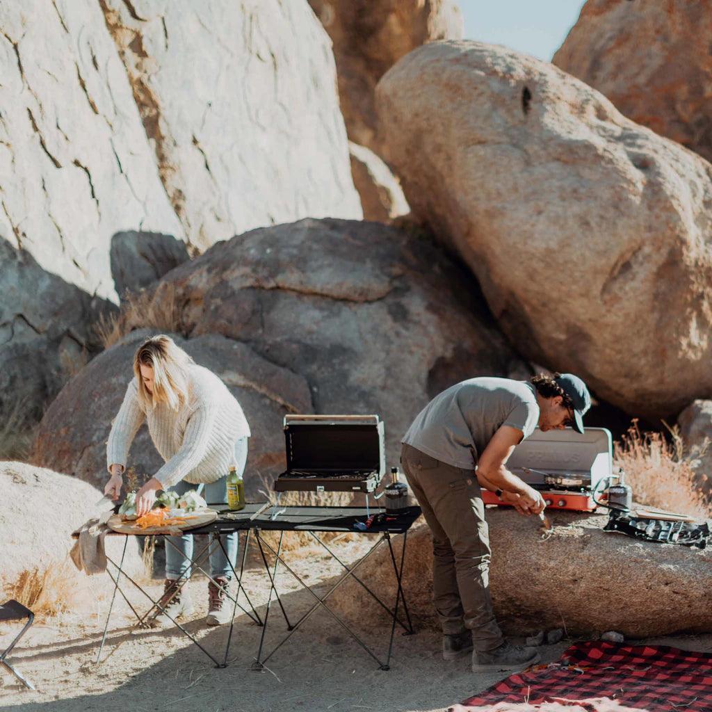 Outdoor camping scene with people using Primus Kuchoma portable gas camp grill on rocky terrain