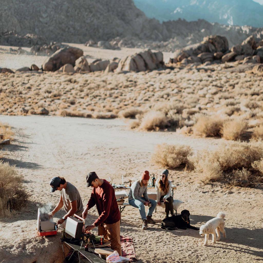 Group camping in desert with Primus Kuchoma portable gas camp grill, cooking outdoors.