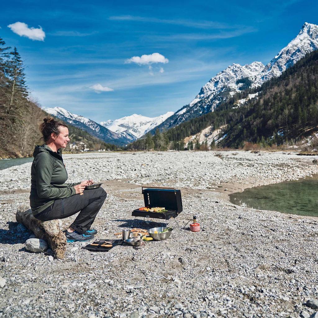 Woman using Primus Kuchoma portable gas camp grill by a mountain river outdoors