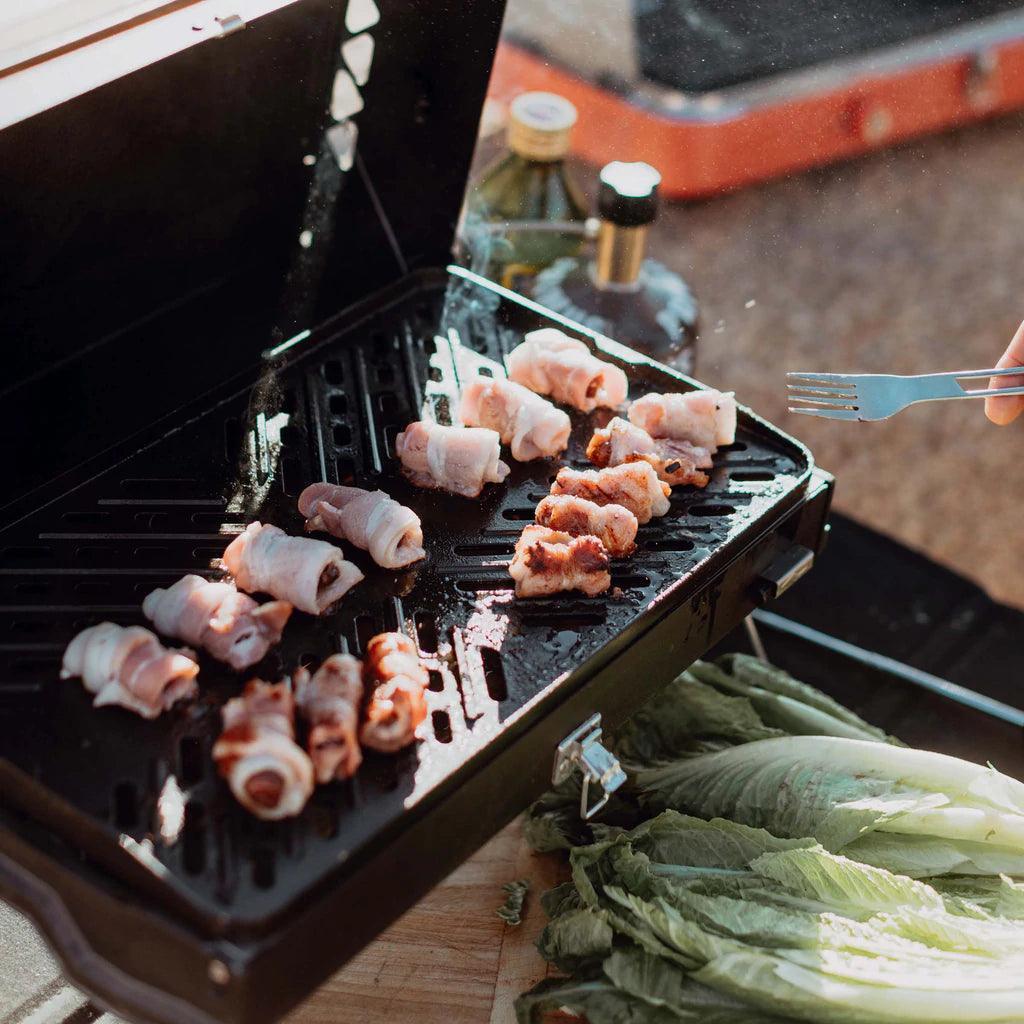Primus Kuchoma portable gas camp grill cooking bacon-wrapped food outdoors with lettuce nearby