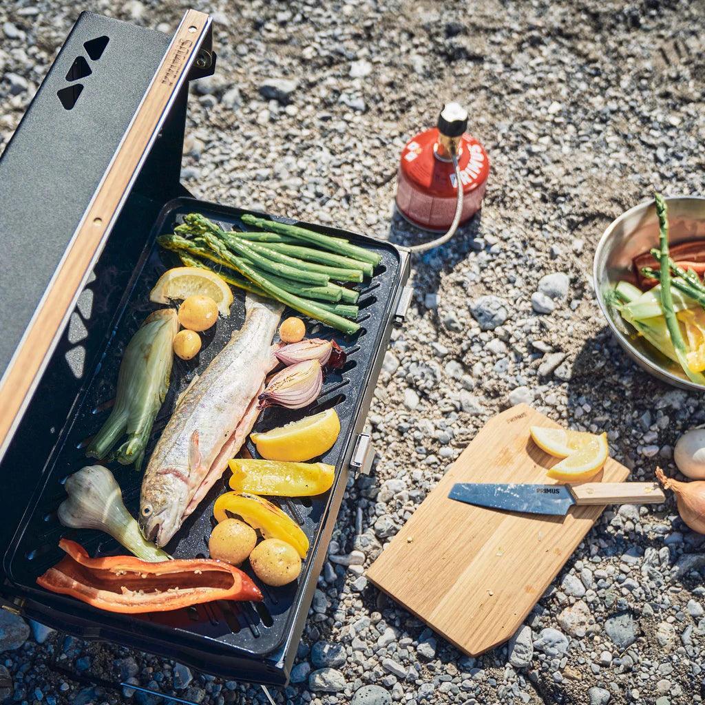 Primus Kuchoma portable gas camp grill cooking fish and vegetables outdoors on gravel