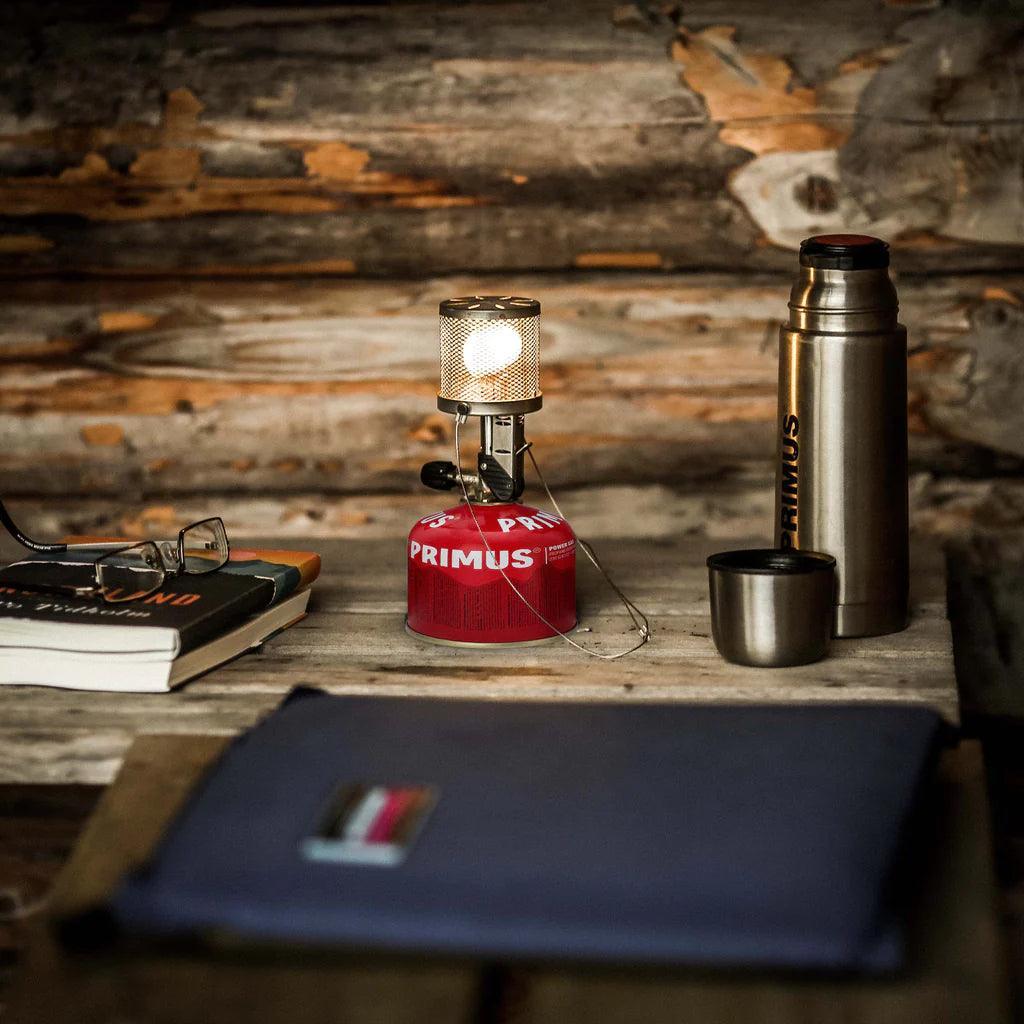 Primus Micron lantern with steel mesh on a rustic wooden table beside a thermos, cup, and books