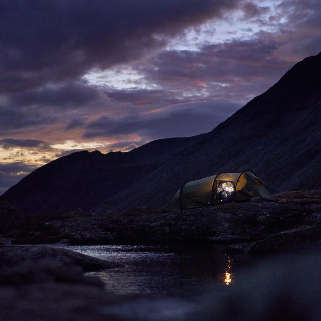 Glowing tent illuminated at dusk in mountain wilderness near water, outdoor camping scene