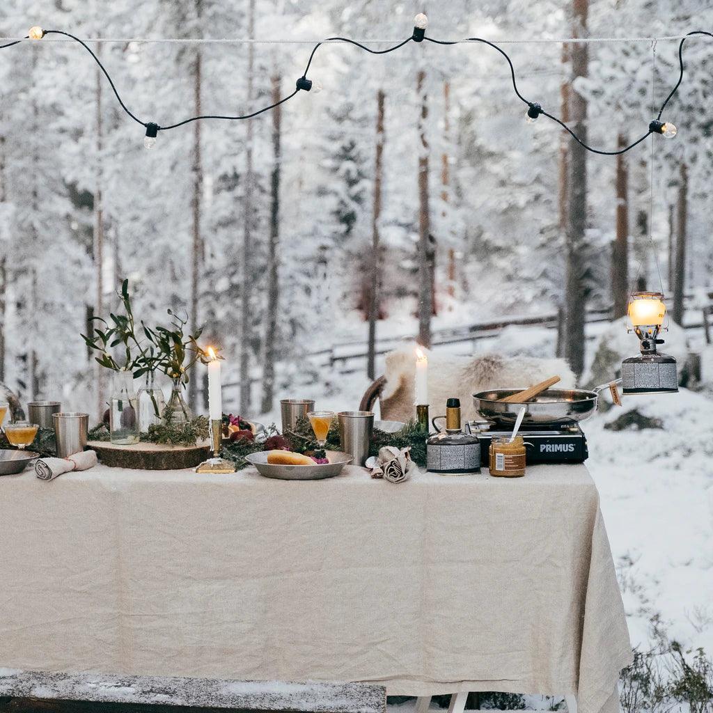 Primus Moja single burner camp stove on snowy outdoor table with food, lantern, and string lights