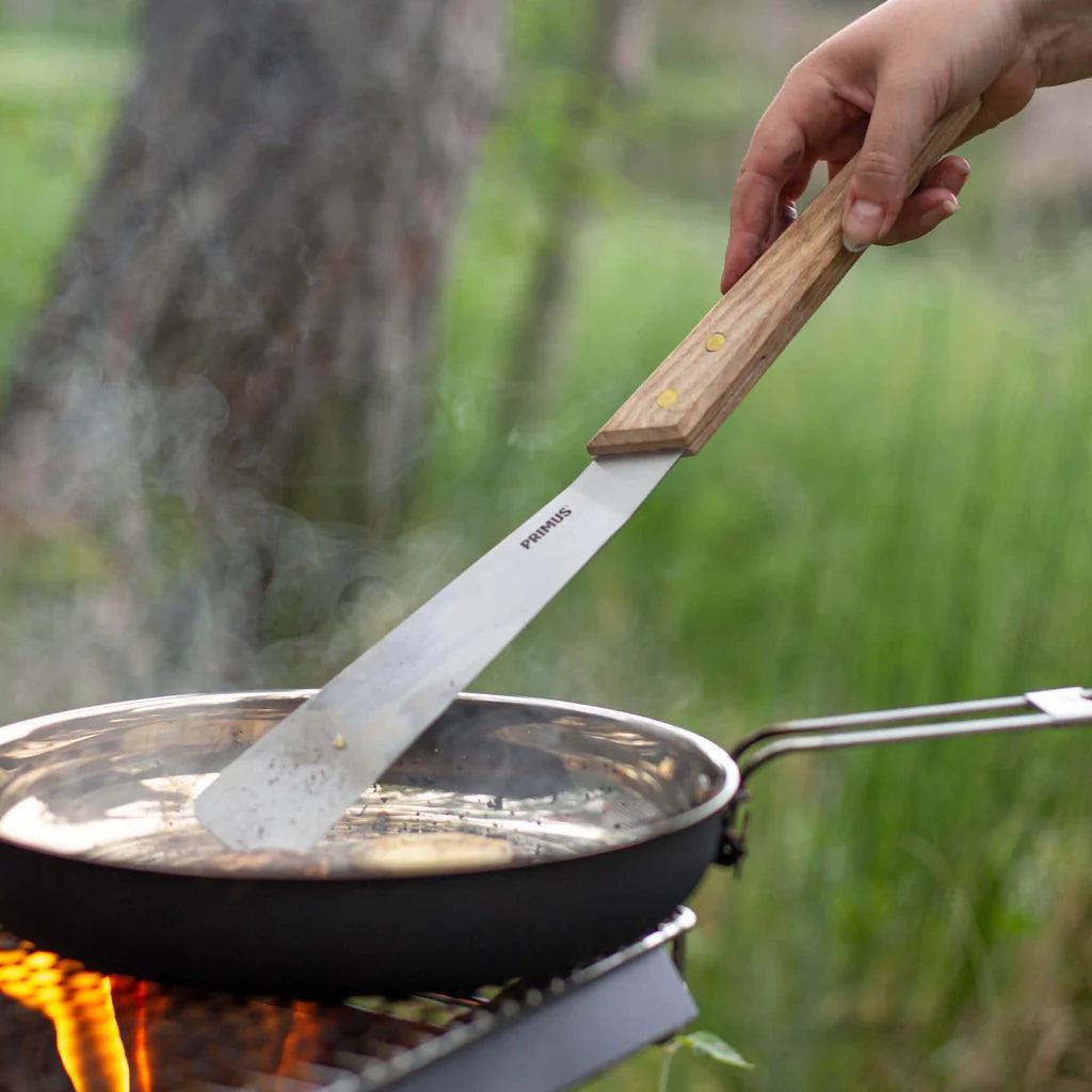 Hand using Primus open fire spatula on frying pan over campfire outdoors
