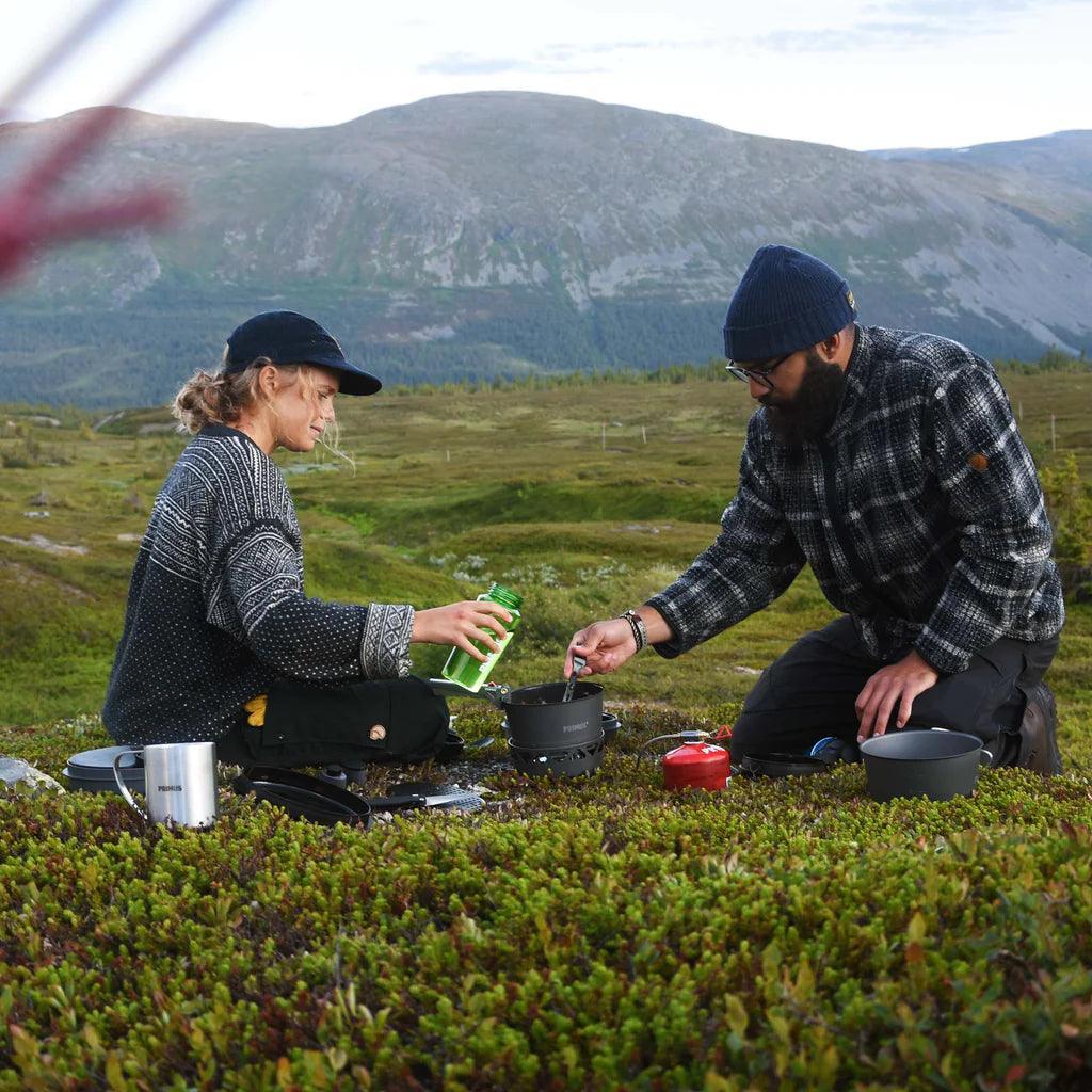 Two people using a Primus PrimeTech stove outdoors in a mountain landscape