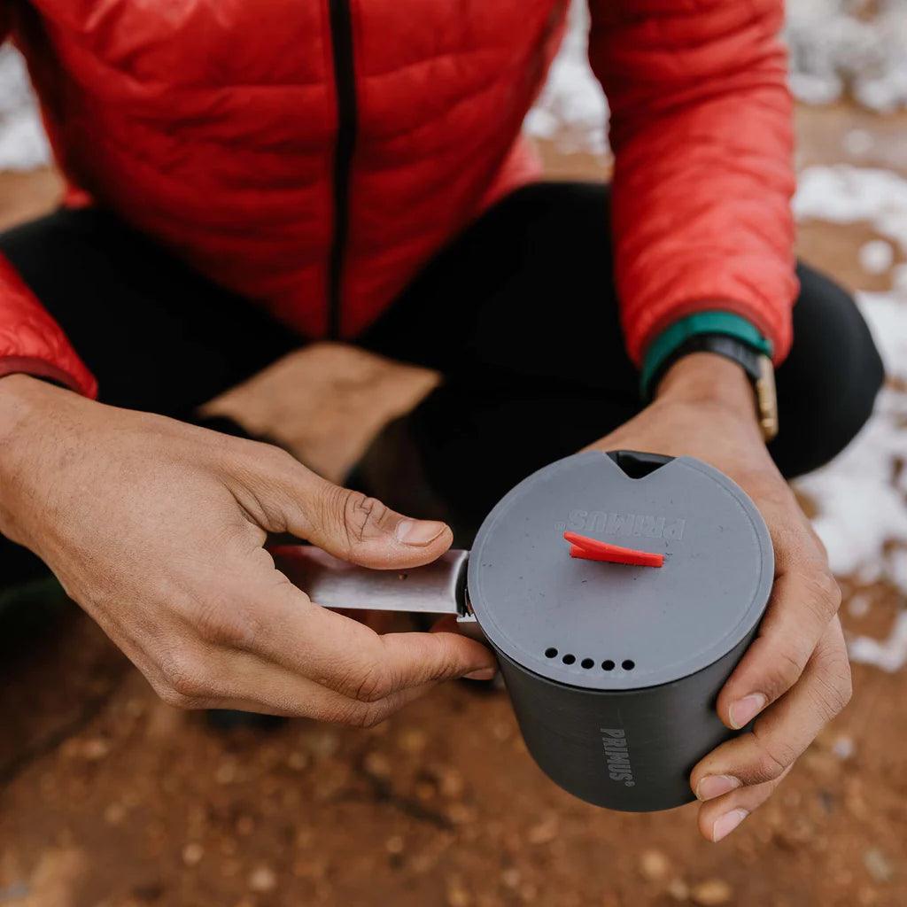 Person holding Primus Trek pot outdoors on dirt ground, wearing red jacket