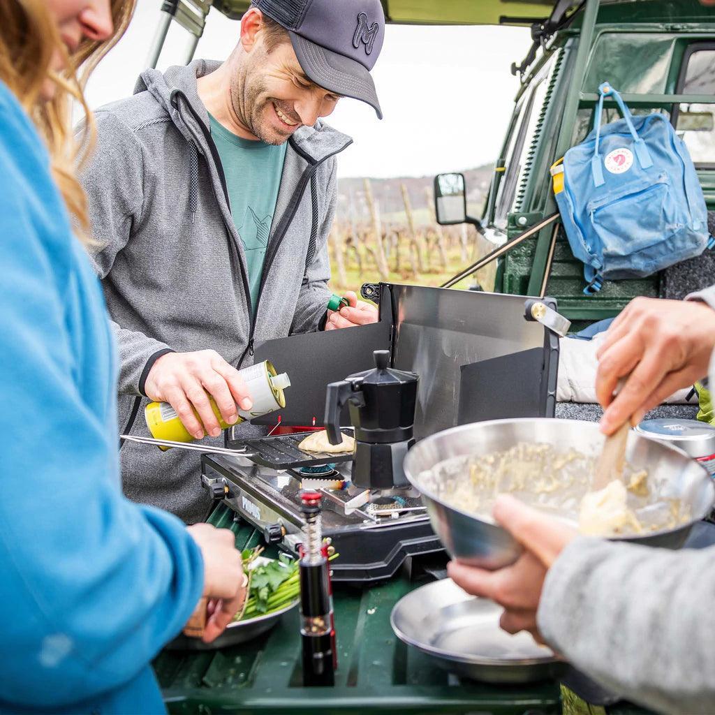 People cooking outdoors with a Primus Tupike stove, mixing ingredients and grilling food at a campsite.