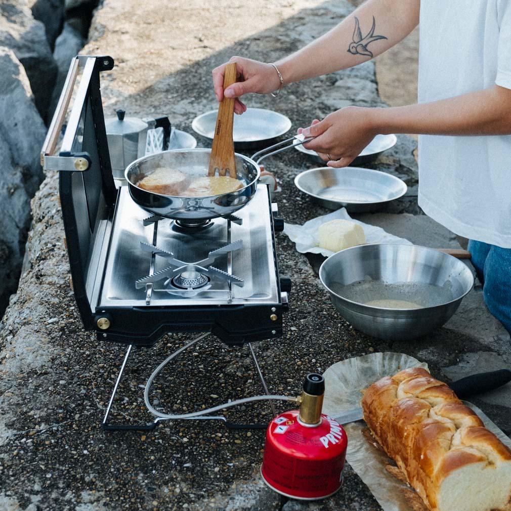 Person cooking with Primus Tupike camping stove outdoors, bread and mixing bowls nearby