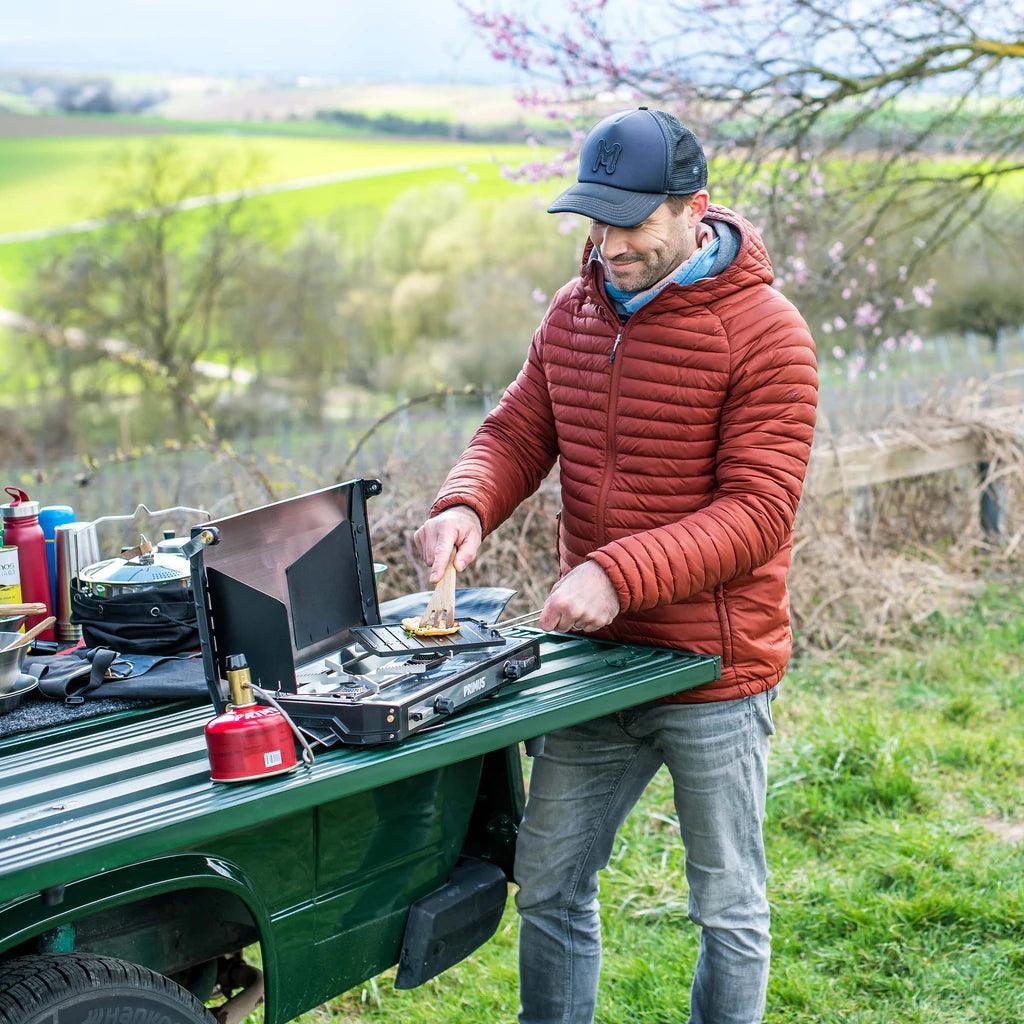 Man cooking outdoors on a Primus Tupike stove set up on a vehicle tailgate in nature