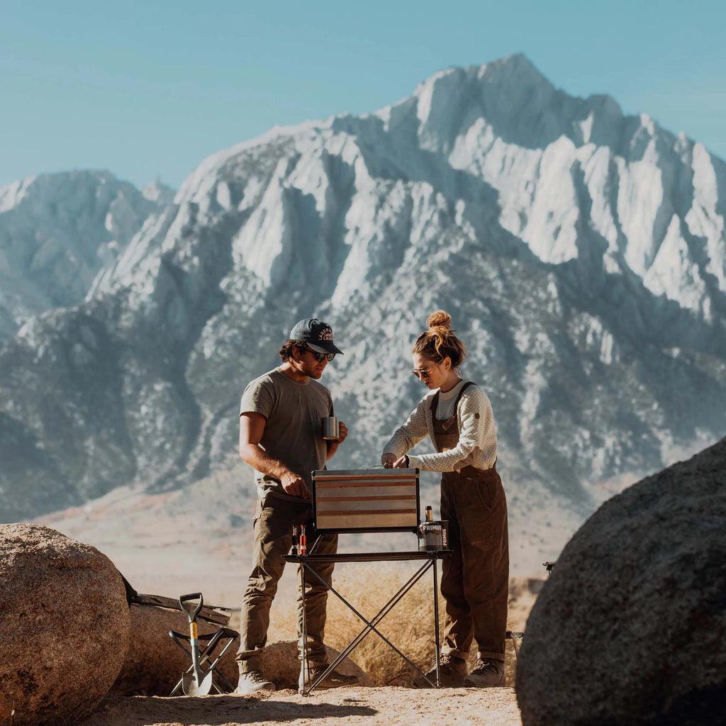 Two people using a Primus Tupike stove outdoors with mountains in the background