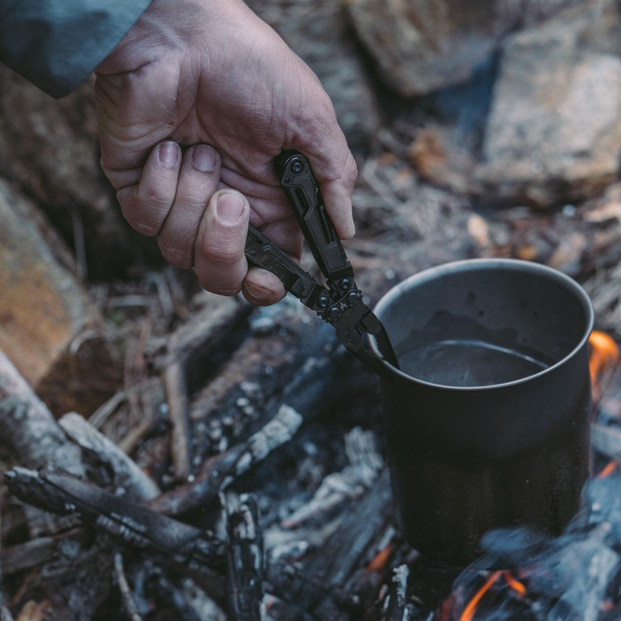 Hand using a black multi-tool plier to hold a metal cup over a campfire outdoors