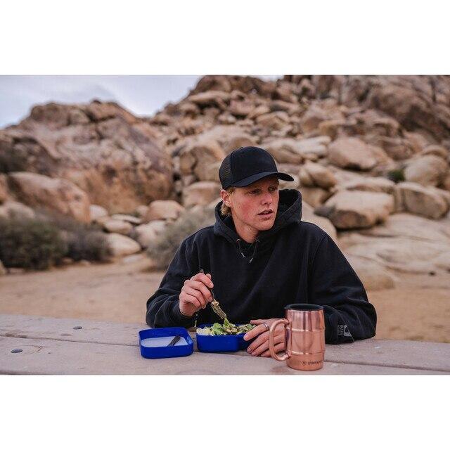 Camper eating meal at outdoor table with blue camping dishware and copper mug, rocky desert background