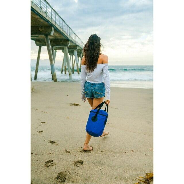 Woman with blue outdoor trail bucket walks barefoot on sandy beach near pier