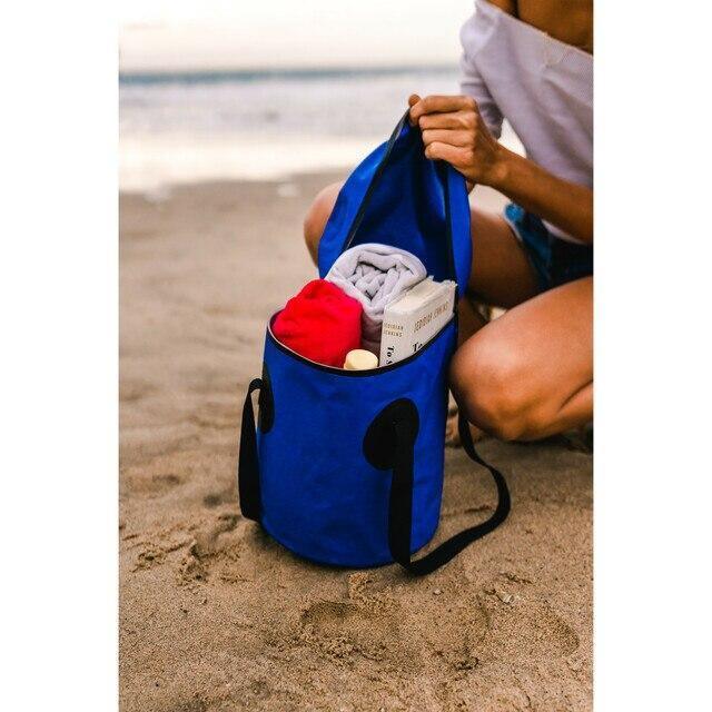 Blue outdoor trail bucket with lid on sandy beach, filled with towels and sunscreen