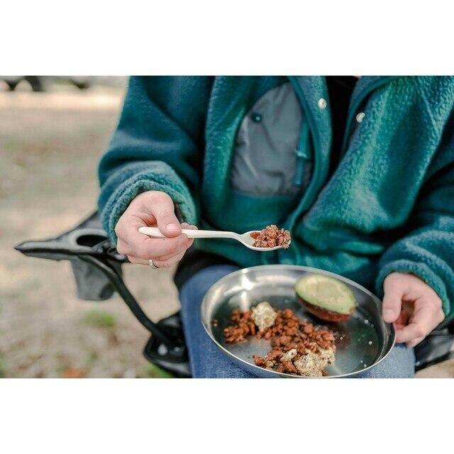 Person using camping cutlery to eat from a metal plate outdoors, camp meal and gear visible