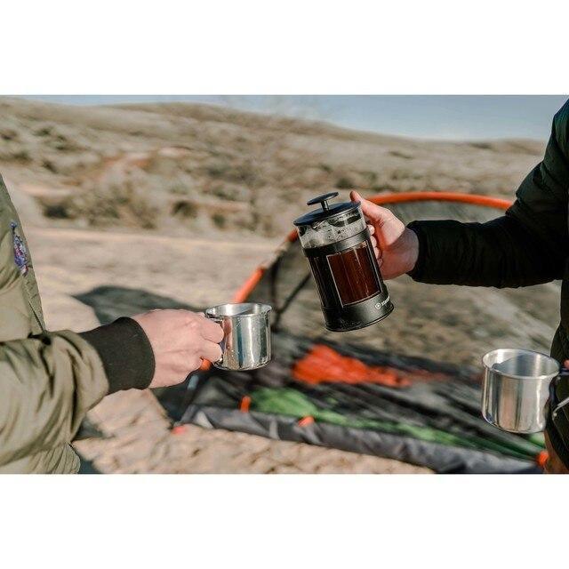 Two people with aluminum drink cups and a French press outdoors near a camping tent
