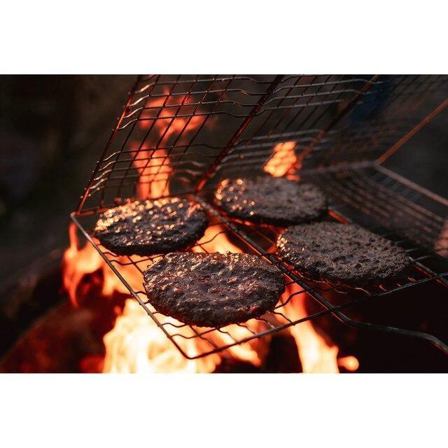 Four burger patties grilling on a metal campfire broiler over open flames outdoors