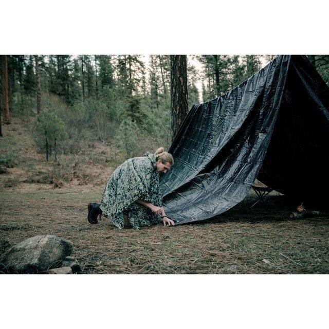 Person in green camo hooded poncho setting up black tarp tent in forest camping area