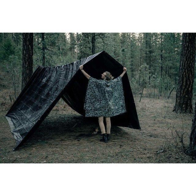 Person in camo hooded poncho setting up black tarp shelter in pine forest outdoors