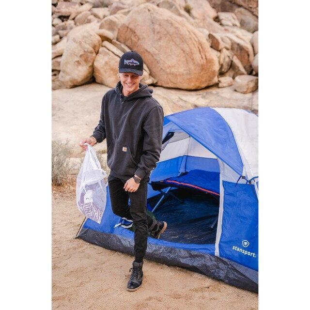 Man holding mesh laundry bag by blue Stansport tent outdoors on sandy ground